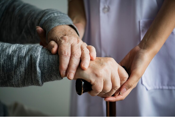 Foto com close em mãos de uma pessoa idosa sendo apoiada pelas mãos de uma cuidadora.
