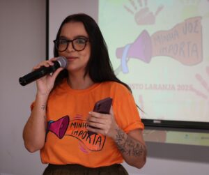 Mulher segurando microfone e celular durante palestra do projeto Minha Voz Importa, vestindo camiseta laranja da campanha Agosto Laranja 2025, com foco em conscientização sobre Esclerose Múltipla.