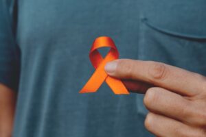 Foto de homem, foco na mão segurando um laço laranja que simboliza o Agosto Laranja. Agosto Laranja e Branco é o mês de conscientização sobre duas doenças graves: a Esclerose Múltipla e o Câncer de Pulmão.