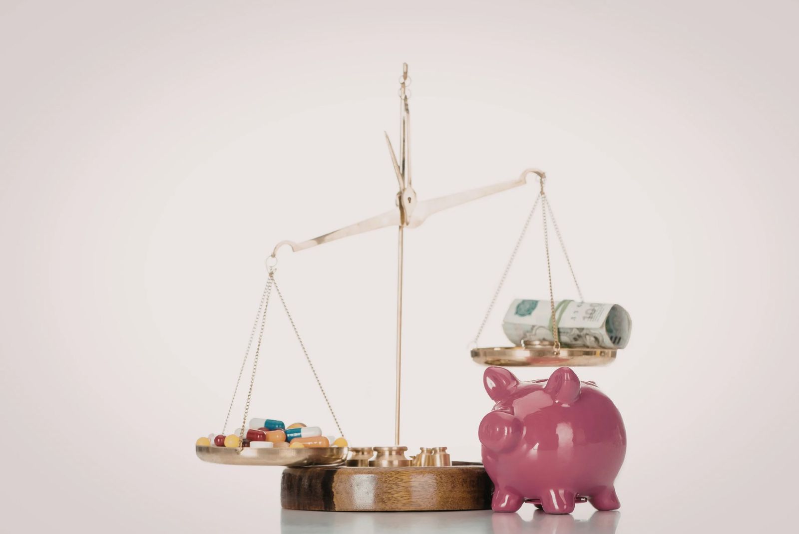 Balança de um lado com medicamentos e o outro com uma nota de dinheiro e um cofre de porquinho